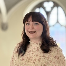 A smiling person with shoulder-length auburn hair and bangs, wearing a light-colored, patterned blouse. They are indoors, standing in front of softly blurred arched windows that let in natural light, creating a warm and airy atmosphere.